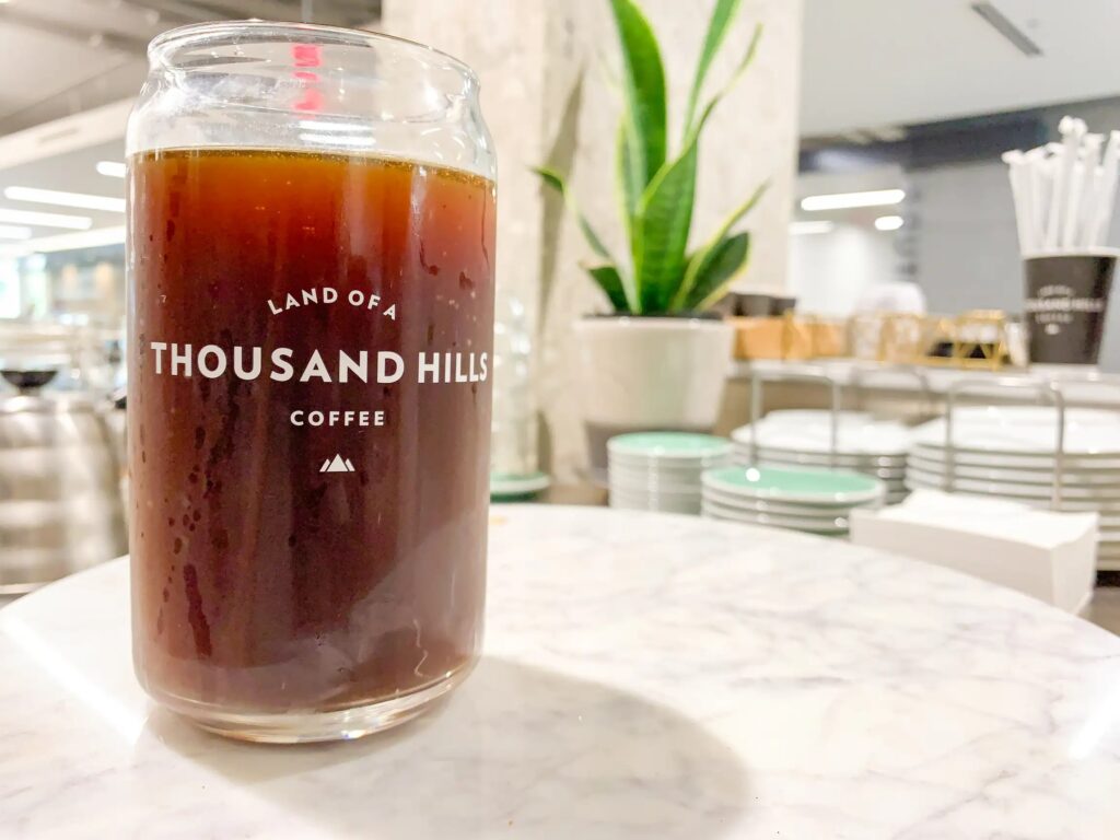 A glass of dark coffee branded "Land of a Thousand Hills" sits on a marble surface. Background features a leafy plant and neatly stacked dishes, conveying a modern café ambiance.
