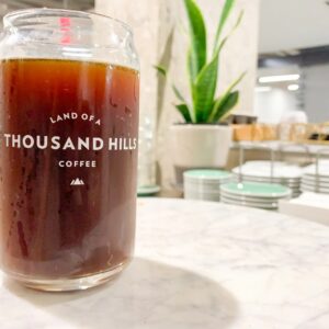 A glass of dark coffee branded "Land of a Thousand Hills" sits on a marble surface. Background features a leafy plant and neatly stacked dishes, conveying a modern café ambiance.